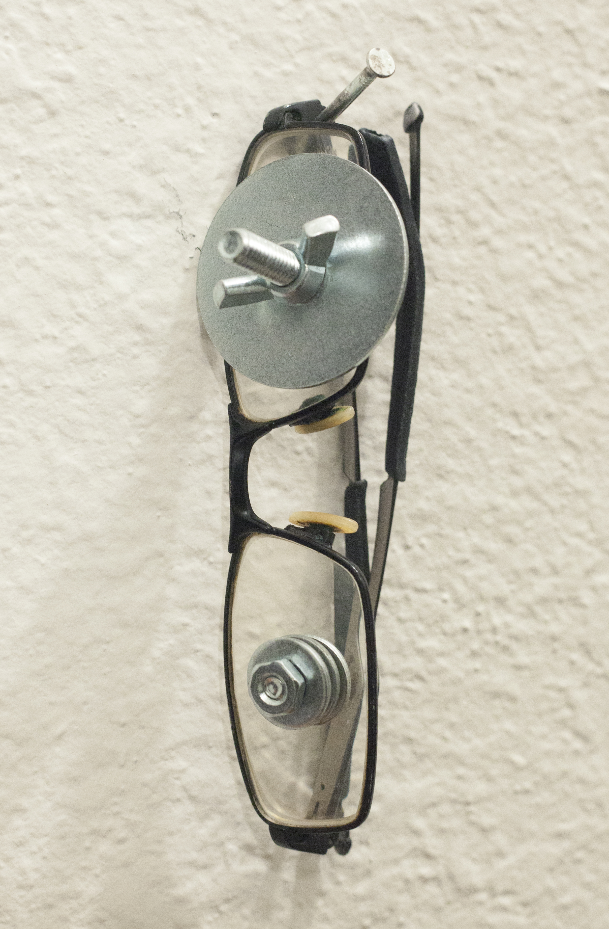 A pair of glasses with screws and washers through the lenses hung on a white wall
